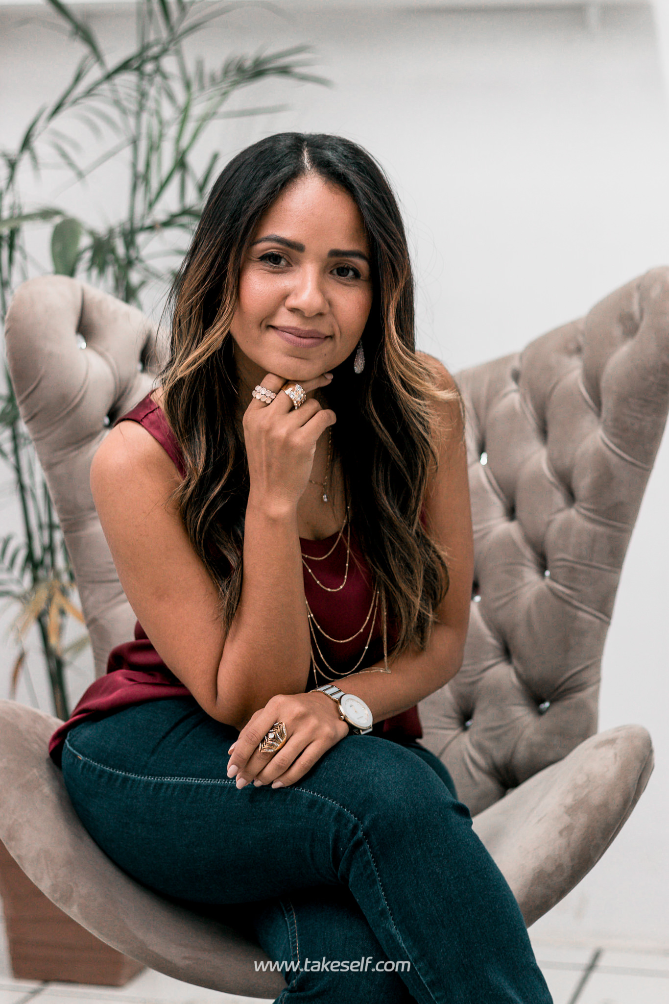 Mulher sorrindo sentada em uma cadeira, usando joias e roupas casuais.