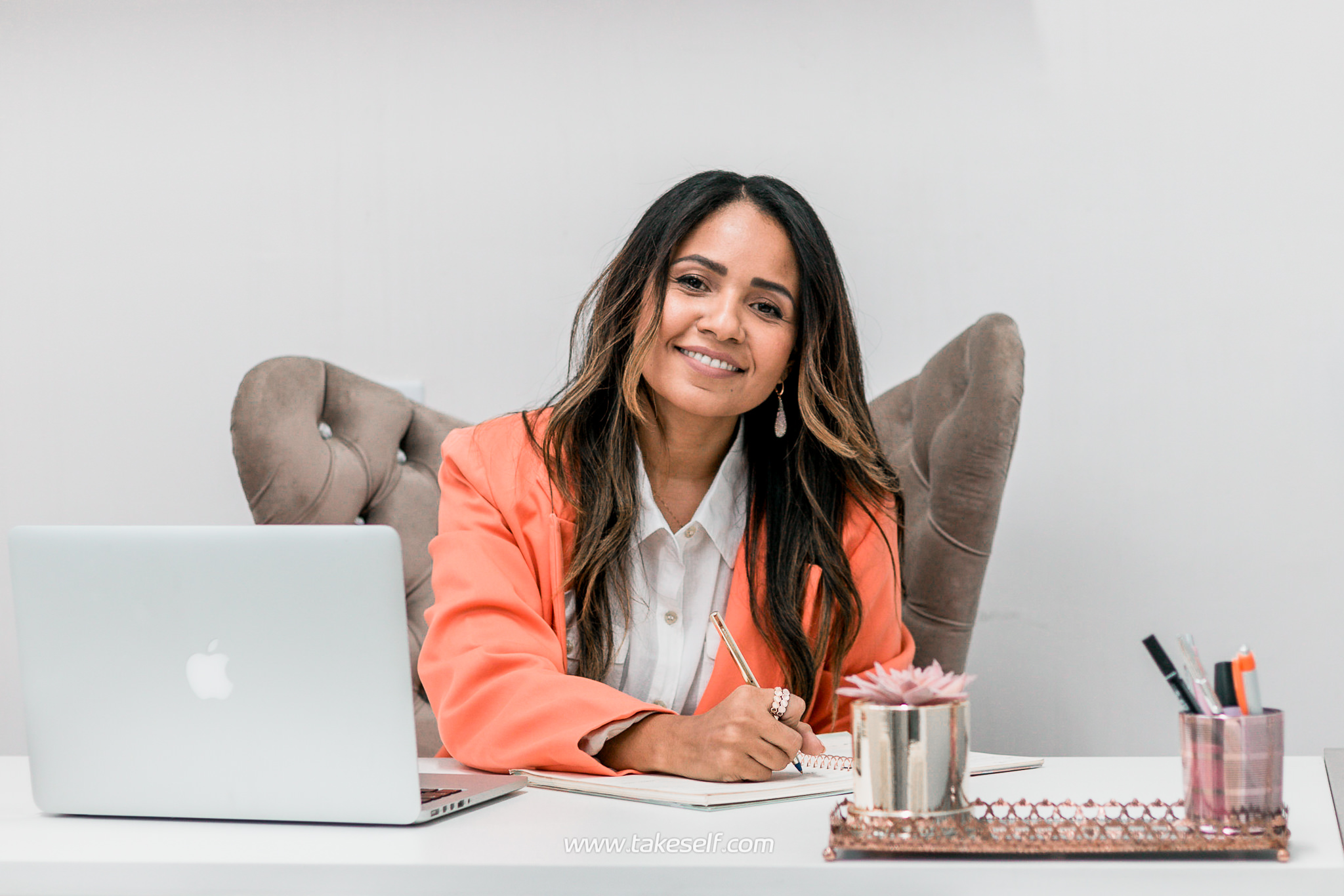 Mulher sorrindo em sua mesa com laptop, escrevendo em um caderno.