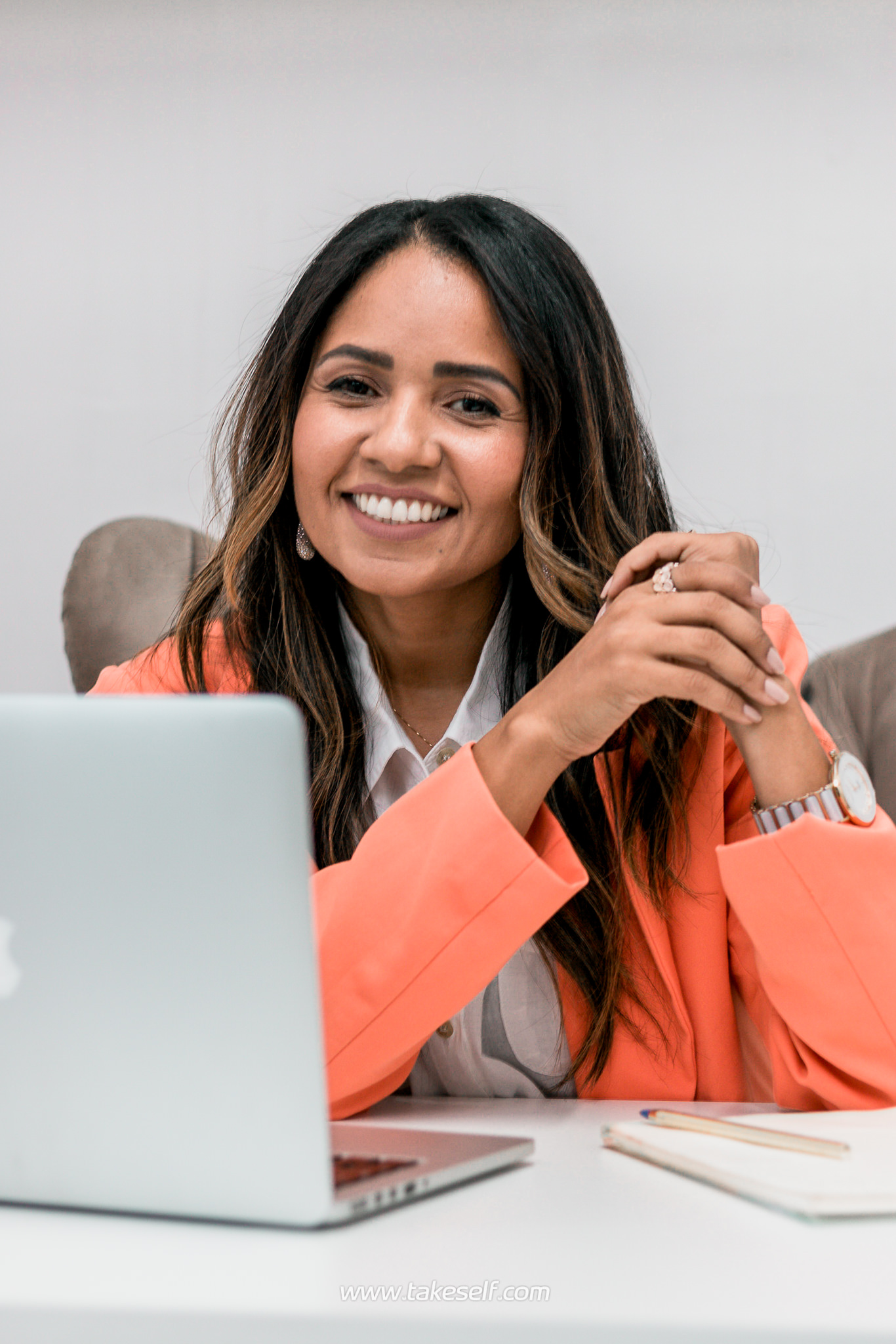 Mulher sorrindo sentada à mesa com um laptop, usando um blazer laranja.